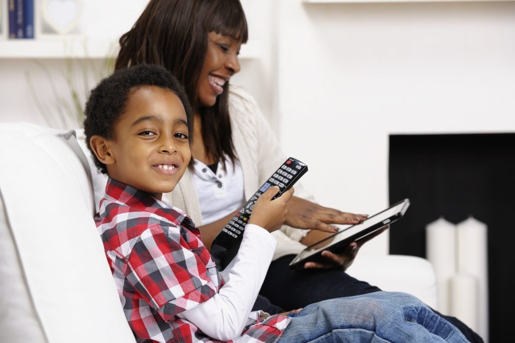 Child holding TV remote control whilst an older woman is looking into a tablet next to him